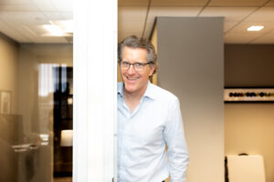 A smiling man in a light blue button-down shirt peeks around a white door frame in an office.